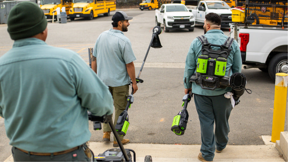 Three men in green uniforms with landscaping power tools walking on a street