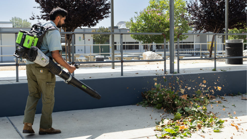 Person using a backpack leaf blower in an outdoor setting with trees and buildings in the background
